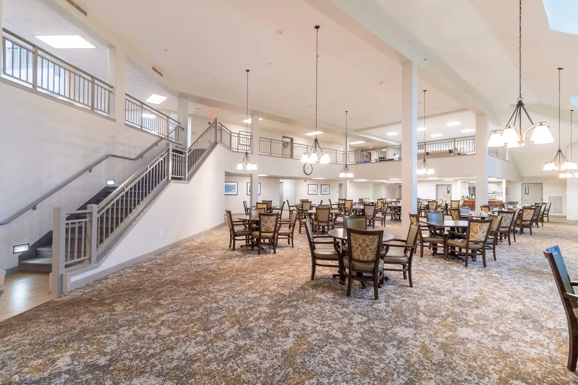 Spacious dining area with multiple round tables and chairs arranged on a patterned carpet. The room features high ceilings with hanging light fixtures and a staircase leading to an upper level with railings. The walls are light-colored, and there are framed pictures and a clock on the far wall.