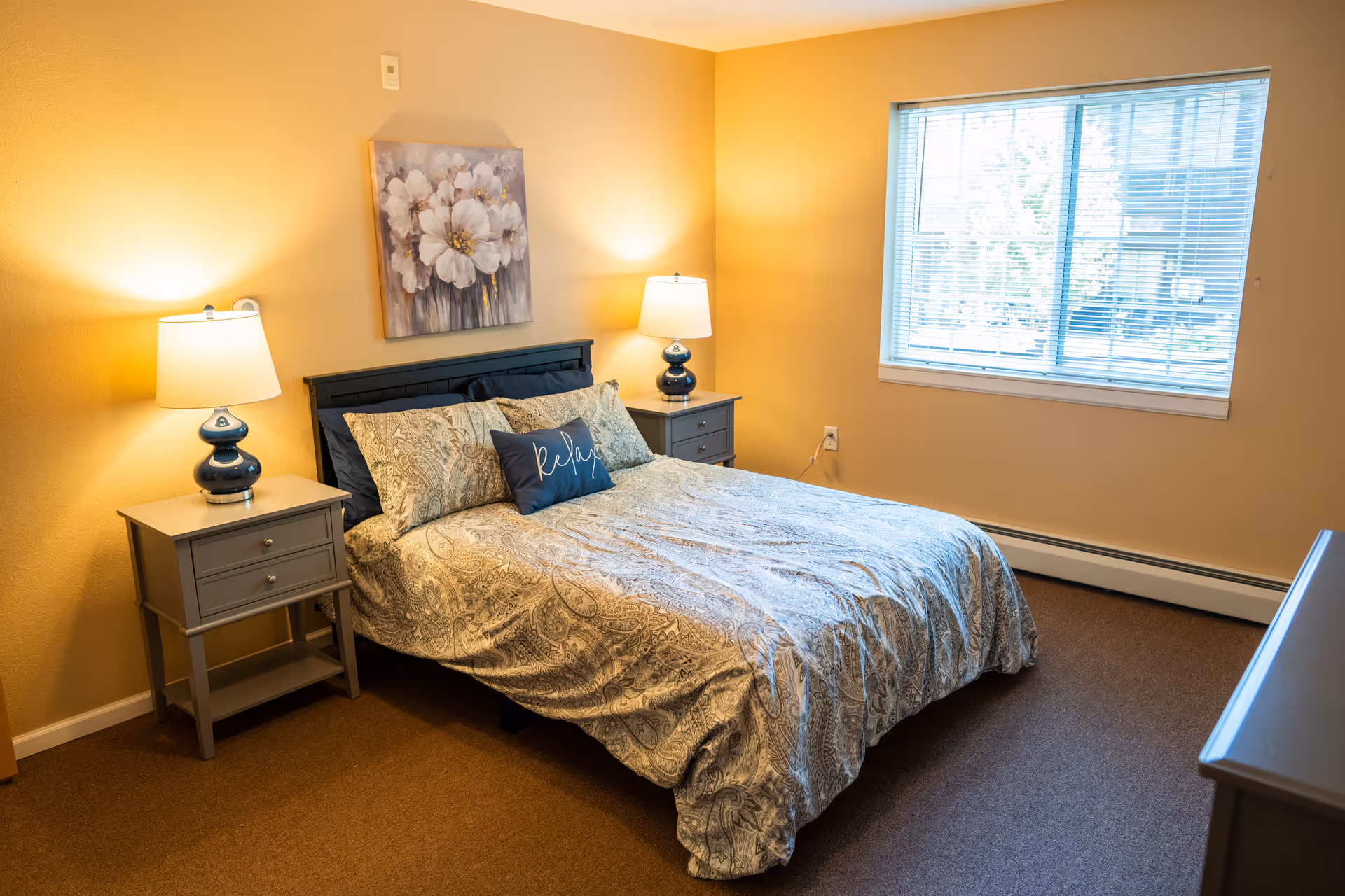 A cozy bedroom with a double bed covered in patterned bedding and a decorative pillow that says 'relax'. There are two matching bedside tables with blue lamps on each side of the bed. A painting of white flowers hangs above the bed. A large window with blinds lets in natural light, and the walls are painted a warm beige color.