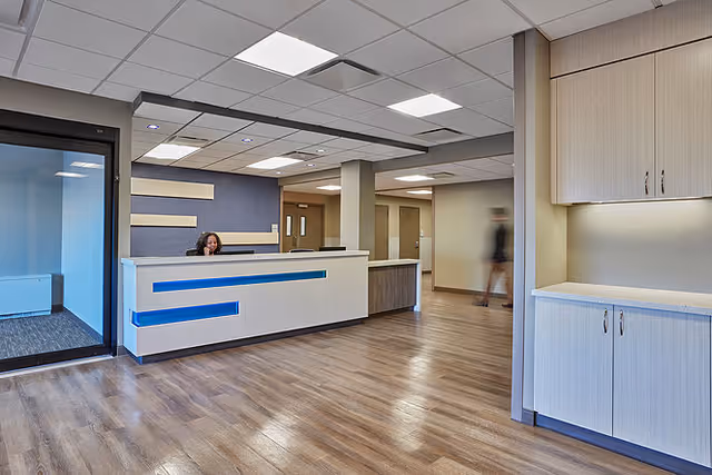 Reception area with a front desk, hardwood floors, ceiling panels, and a staff member seated behind the desk.