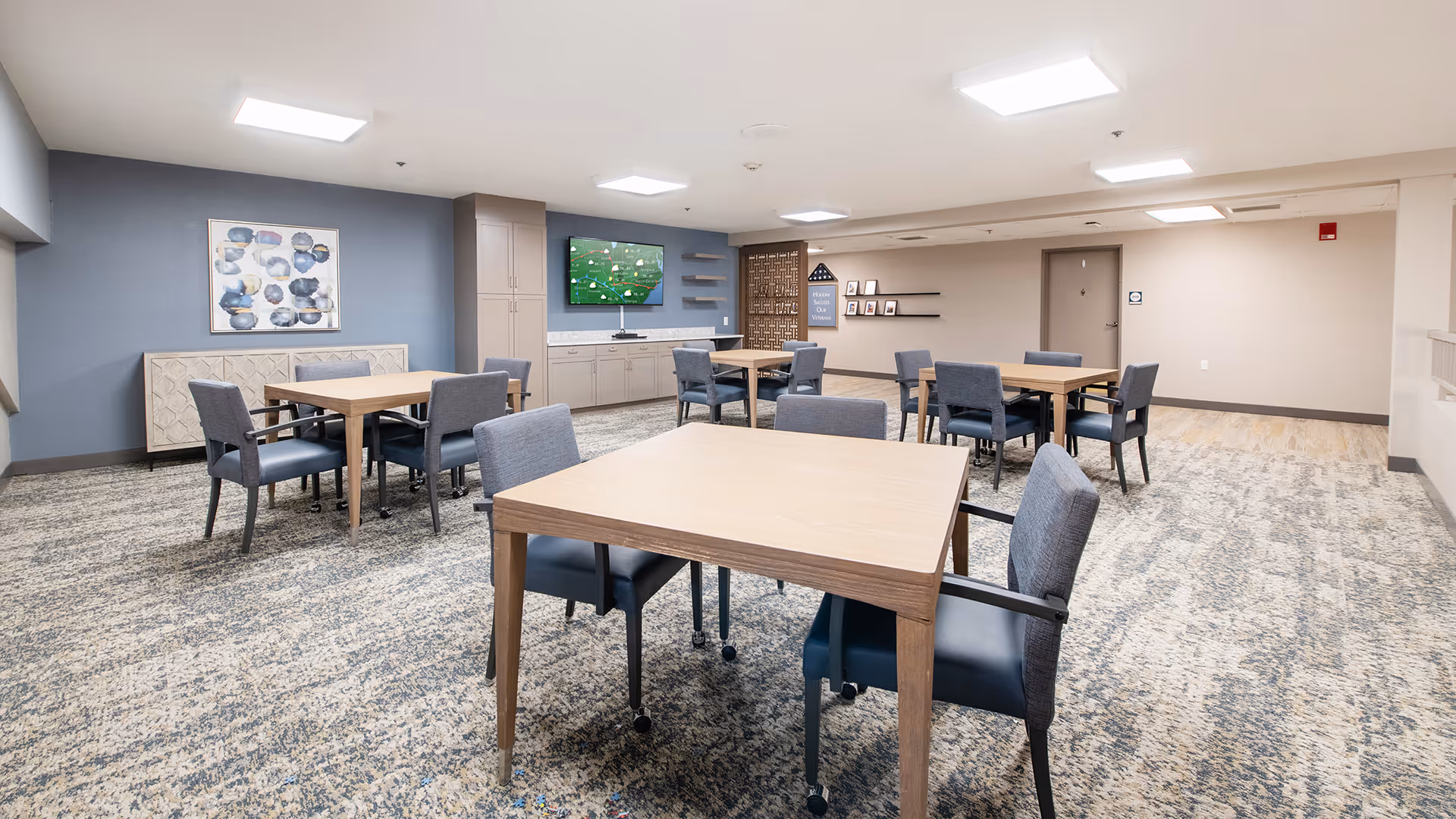 A spacious room with several square wooden tables and gray upholstered chairs arranged around them. The room has a patterned carpet floor, light-colored walls, and a blue accent wall with a framed abstract painting. There is a flat-screen TV mounted on the wall above a built-in cabinet, and some decorative shelves and framed photos on the far wall.
