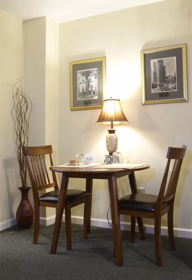 A small wooden dining table with two wooden chairs with black cushions is set against a light-colored wall. On the table, there is a decorative lamp, a glass, a coffee cup, and some small items. Two framed black and white pictures hang on the wall above the table. A tall decorative vase with dried branches is placed in the corner beside the table.
