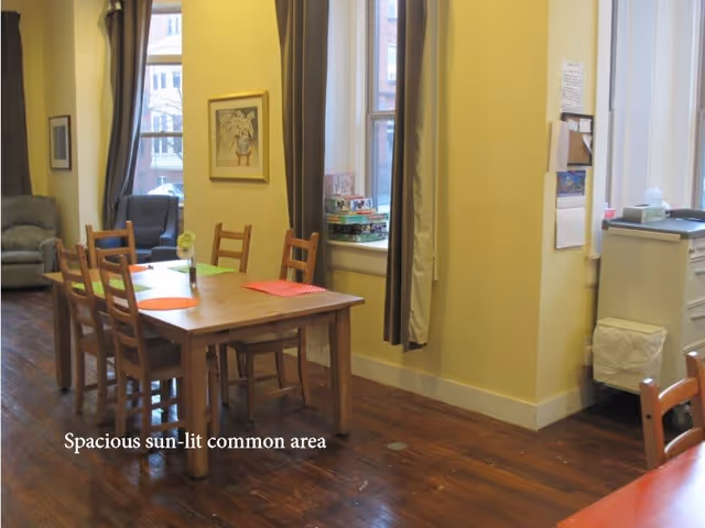 A spacious sun-lit common area with a wooden table and six chairs, colorful placemats on the table, a small vase with flowers, large windows with curtains letting in natural light, a few board games stacked on the windowsill, and comfortable armchairs in the background.
