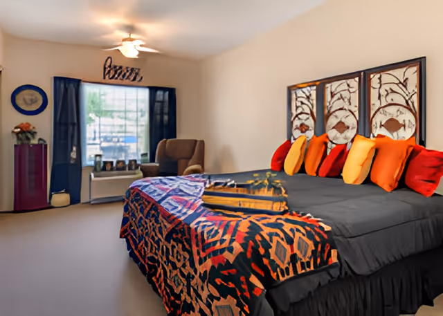 A bedroom with a large bed covered in a black and colorful patterned bedspread and multiple orange and red pillows. Behind the bed is a decorative headboard with a nature-inspired design. There is a window with dark curtains, a brown armchair, and a small table with framed photos and a flower vase in the corner.