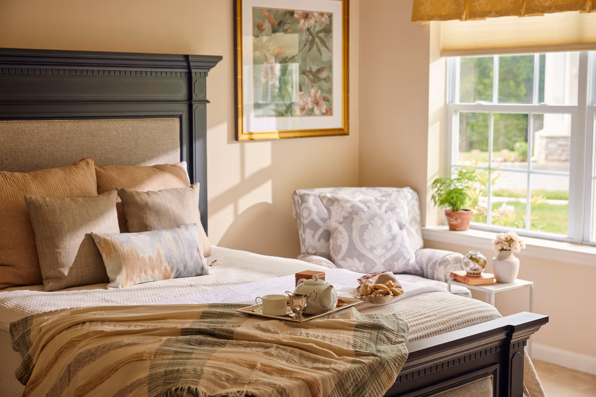 A cozy bedroom with a large bed featuring multiple pillows and a patterned blanket. A tray with a teapot, cup, and croissants is placed on the bed. Next to the bed is a comfortable armchair with a floral pattern, a small side table with a potted plant and a vase of flowers, and a window letting in natural light.