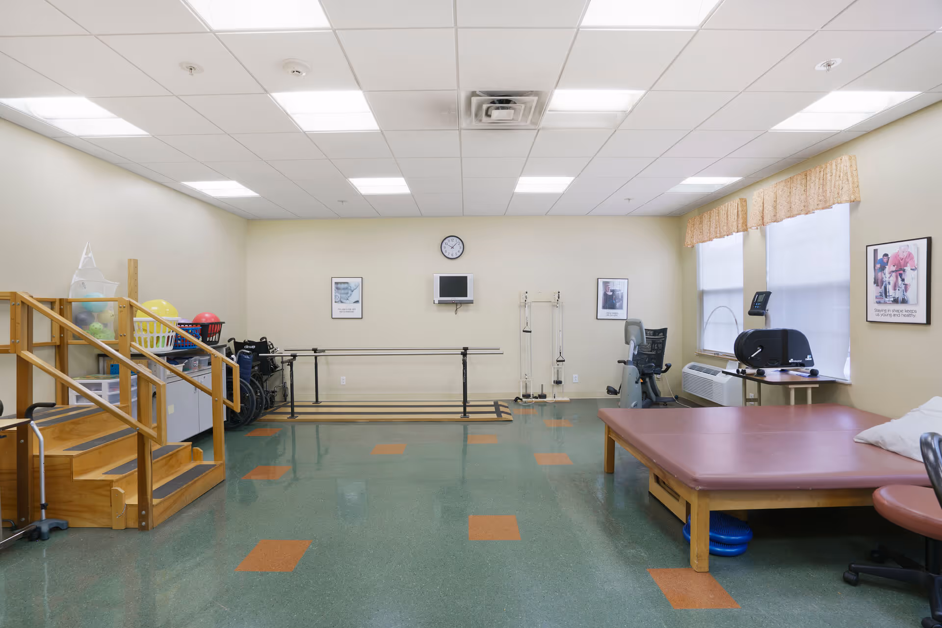 A physical therapy or rehabilitation room with wooden stairs for practice, parallel bars for walking exercises, exercise balls, wheelchairs, a large padded therapy table, and exercise equipment. The room has a green and orange tiled floor, beige walls, fluorescent ceiling lights, and two windows with valances. There are framed pictures on the walls and a clock above a small mounted TV.