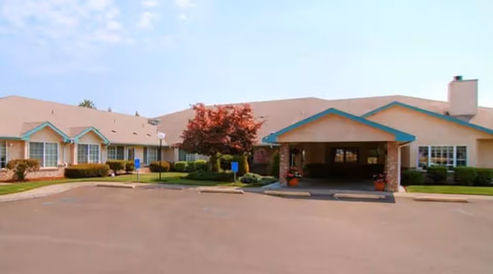 Exterior view of Regency Park Place at Corvallis, showing a single-story building with beige walls and a brown roof. The entrance has a covered porch supported by brick columns, with potted plants on either side. There is a parking lot in front with some handicap parking signs and landscaped bushes and trees around the building.
