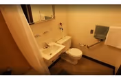 Small bathroom with a wall-mounted sink, toilet, and a shower area with a curtain. A mirror is mounted above the sink, and there is a towel rack with a blue towel and a white towel on the wall. The walls are painted light yellow, and the floor has a brownish carpet or mat.