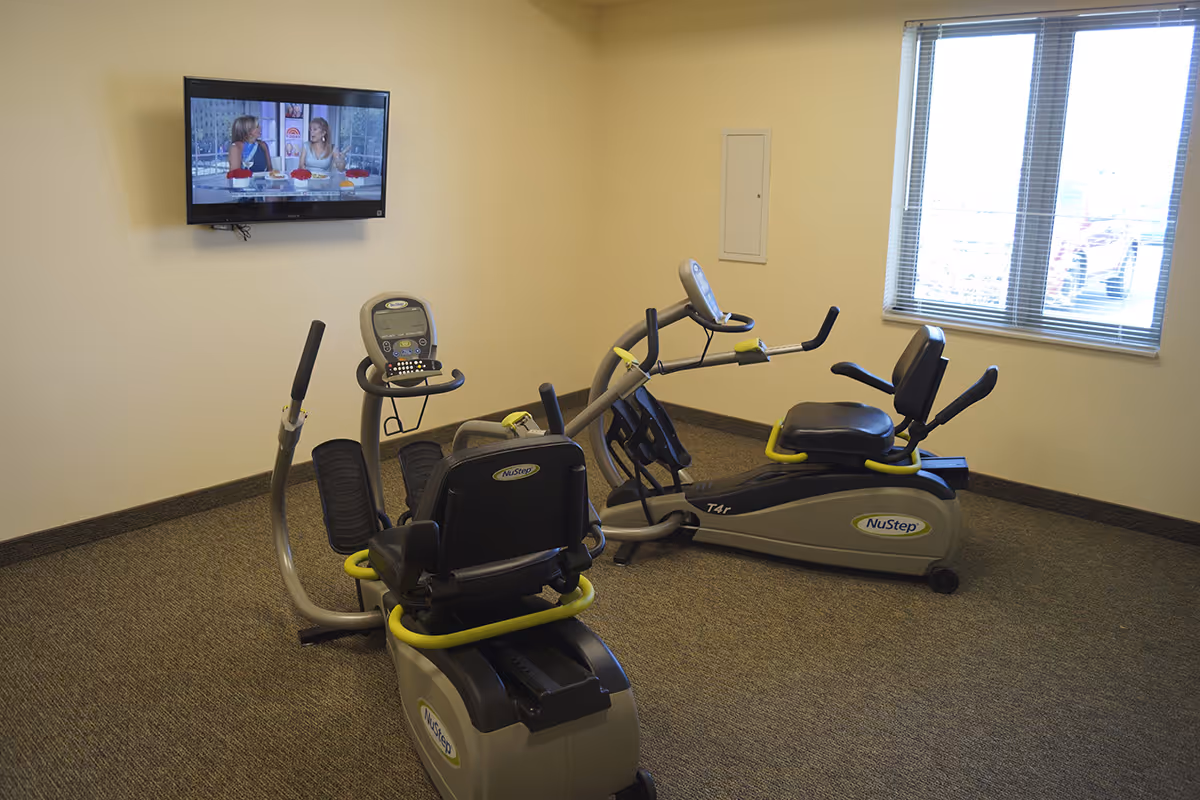 A small exercise room with two NuStep recumbent cross trainers positioned on a carpeted floor. A flat-screen TV is mounted on the wall showing a talk show with two women. There is a window with blinds letting in natural light.