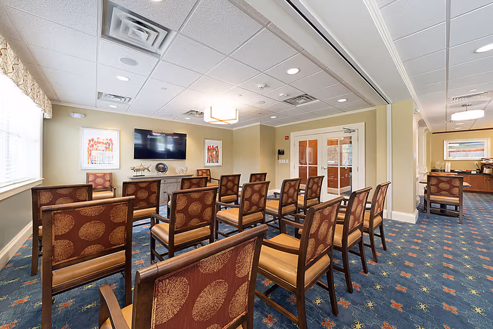 A well-lit meeting or presentation room with rows of wooden chairs featuring patterned upholstery facing a wall-mounted flat screen TV. The room has beige walls, a blue carpet with colorful patterns, two framed artworks on the wall, and a sideboard with decorative items. There are double glass doors on one side and a window with a valance on the other.
