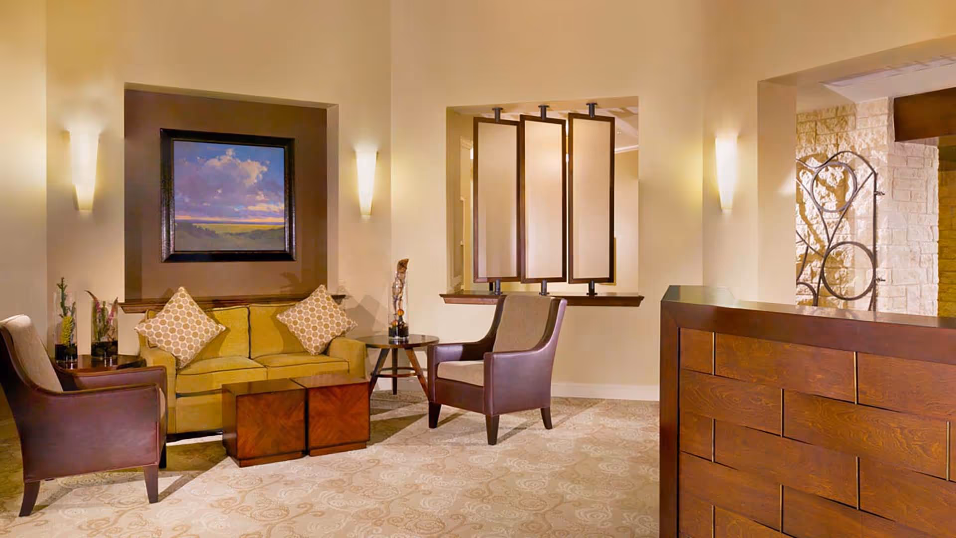 A cozy living room area with a yellow sofa adorned with two patterned pillows, two brown armchairs, a small round wooden table, and two square wooden coffee tables. The walls are painted in warm tones and decorated with a framed landscape painting and modern wall sconces. A wooden reception desk is visible on the right side, and a decorative partition with three panels separates the space.