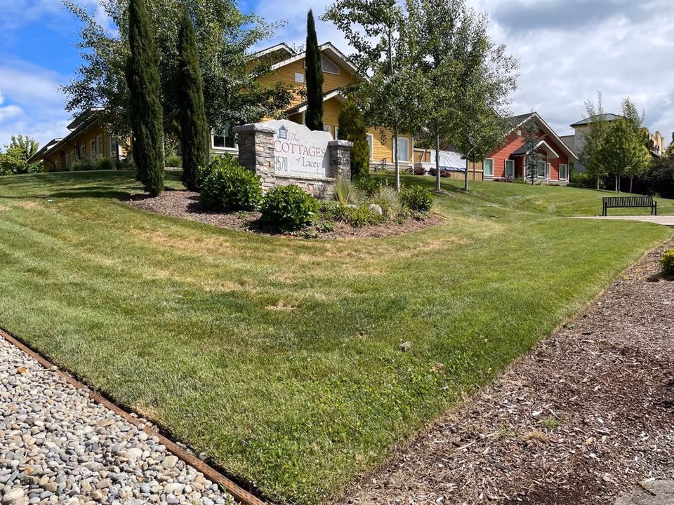 Landscaped lawn and entrance with a stone sign reading 'The Cottages of Lacey' in front of colorful cottage-style buildings.