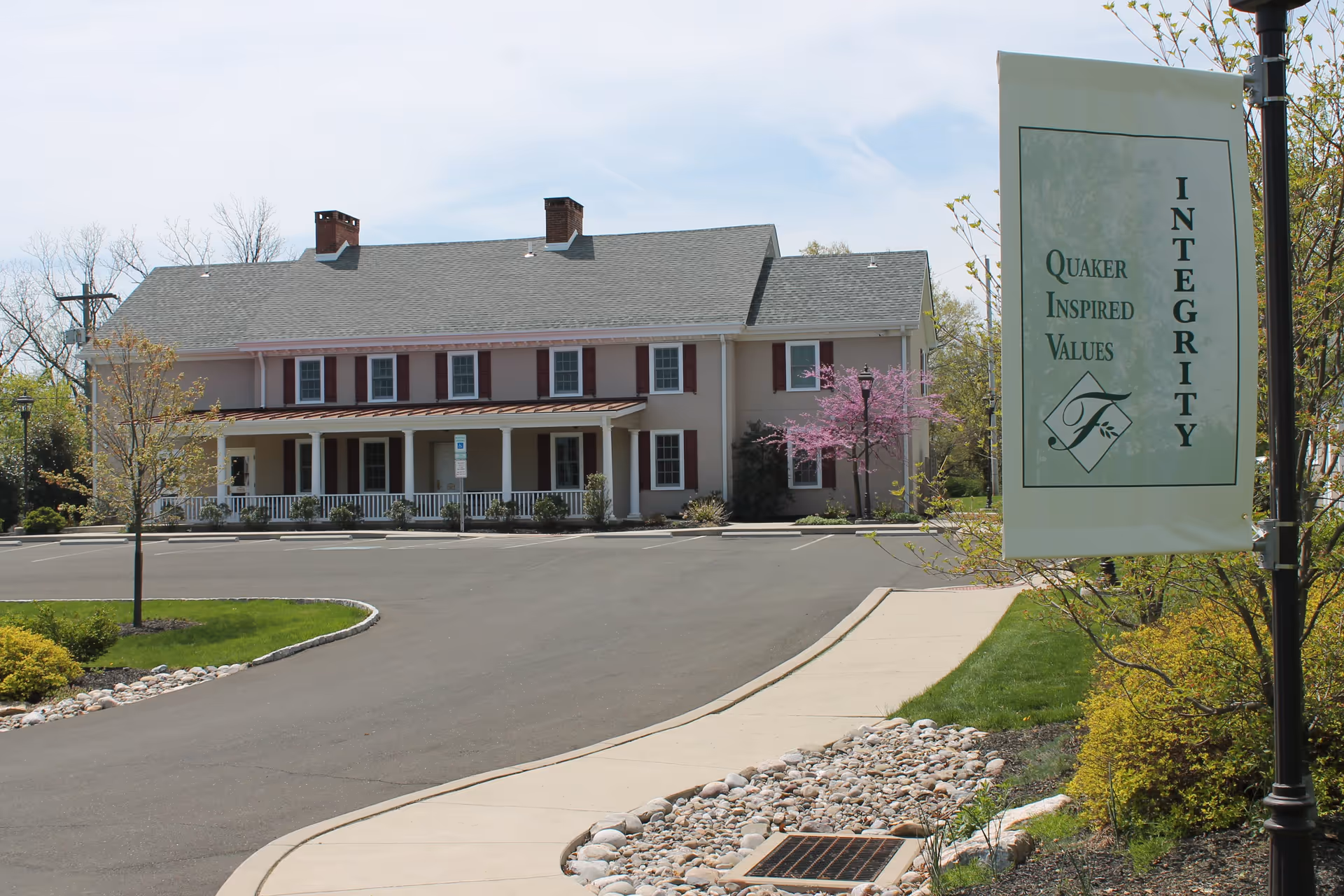 Exterior view of a two-story building with a gray roof and multiple windows, surrounded by a parking lot and landscaped greenery including a small tree with pink blossoms. A sidewalk runs alongside the parking lot, and a sign on a lamp post reads 'Quaker Inspired Values INTEGRITY' with a decorative emblem.