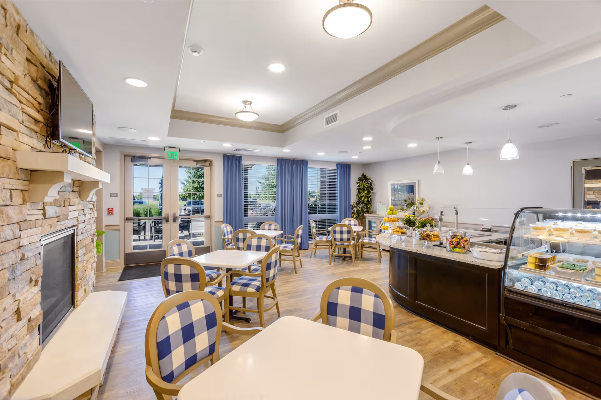 A bright and inviting dining area with several tables and chairs featuring blue and white checkered upholstery. The room has a stone fireplace with a mounted TV above it on the left side. Large windows with blue curtains allow natural light to enter. On the right side, there is a curved counter displaying various food items and desserts in a glass case. The floor is wooden, and the ceiling has recessed lighting and hanging pendant lights.