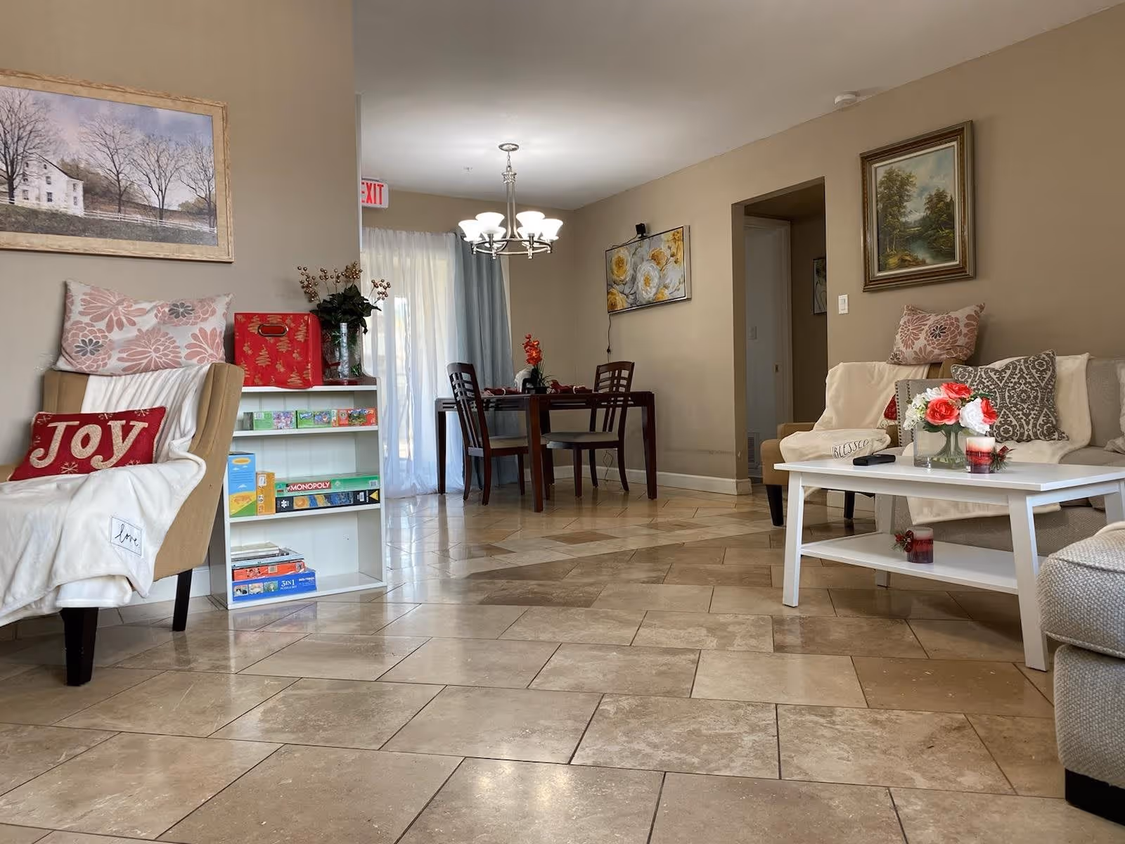 Bright common living area with armchairs and a coffee table with flowers, a small shelf of games, and a dining table in the background.