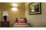 Small single bed with a patterned bedspread against a beige wall, a nightstand with a lamp and items to the left, and a wall-mounted light and framed artwork above.