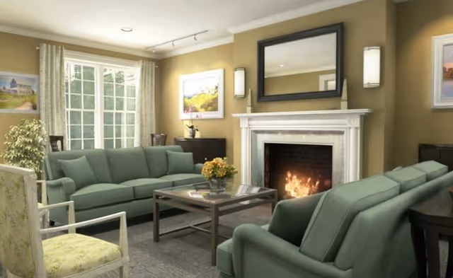 A cozy living room with green upholstered sofas and armchairs arranged around a wooden coffee table. There is a lit fireplace with a large mirror above it, two wall sconces on either side, and framed artwork on the walls. A large window with curtains lets in natural light, and a potted plant is placed near the window.