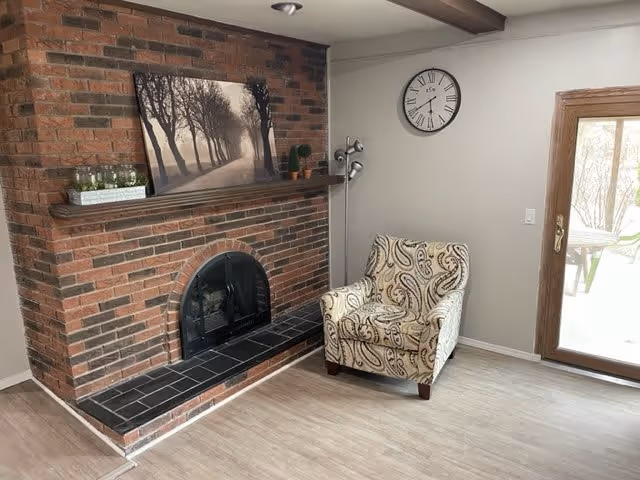 Cozy living room with a brick fireplace, patterned armchair, wall clock, and glass door to the outside.