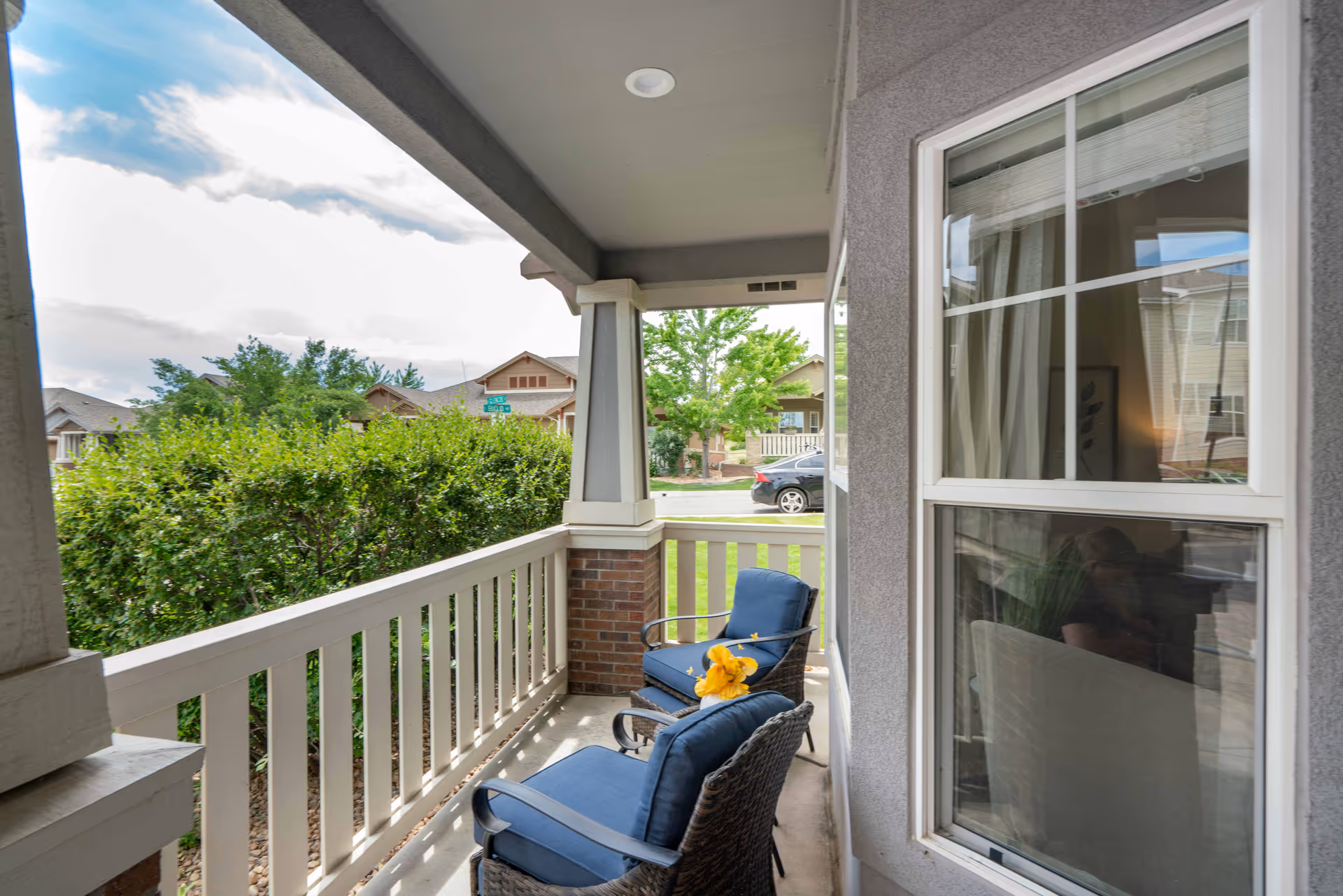 Covered porch area with two blue cushioned chairs, one with a yellow flower on it, overlooking a green hedge and neighborhood houses across the street under a partly cloudy sky.