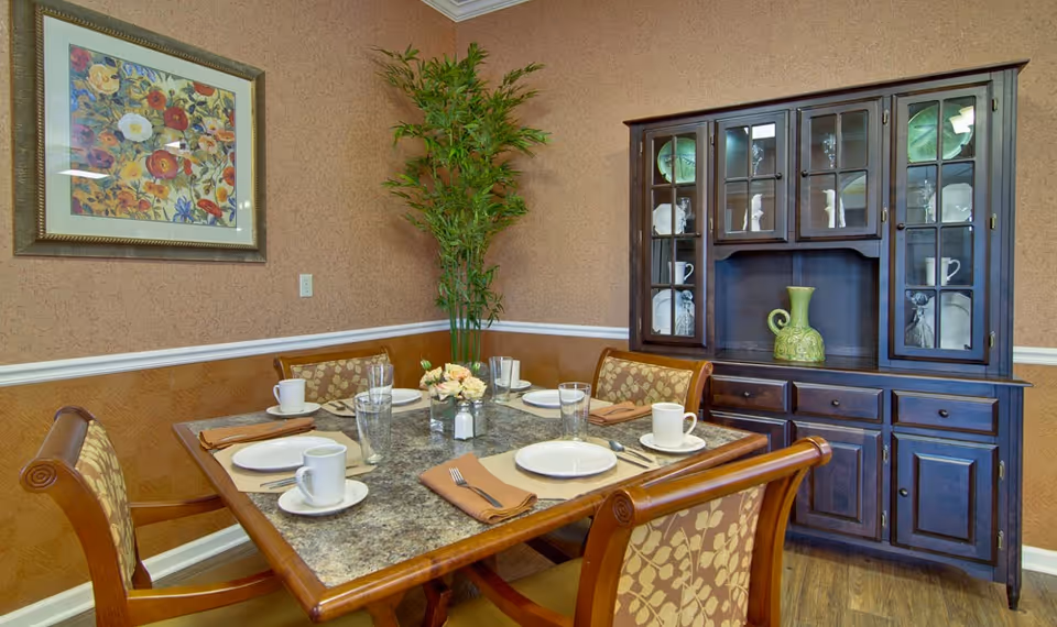 A dining room with a square table set for four people, featuring white plates, cups, glasses, and brown napkins. The room has patterned wallpaper, a framed floral painting on the wall, a tall green plant in the corner, and a dark wooden china cabinet displaying decorative items.