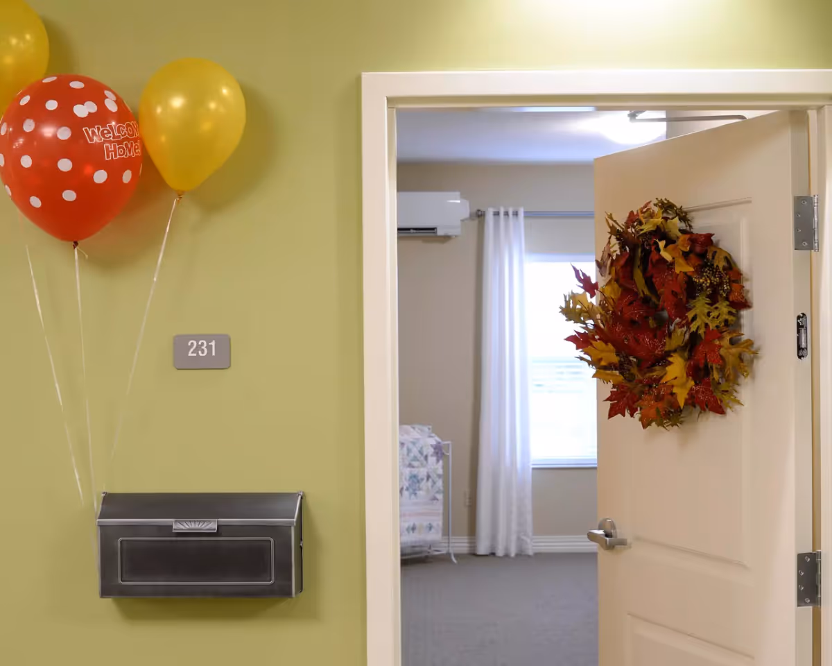 Open door of room 231 in a senior living facility decorated with a fall-themed wreath. Two balloons, one red with white polka dots and the words 'Welcome Home!' and one yellow, are tied to a mailbox mounted on a green wall next to the door. Inside the room, a window with white curtains and a quilt on a bed are visible.