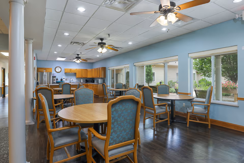 A bright dining area in an assisted living facility with several round wooden tables surrounded by cushioned chairs with wooden frames. The room has large windows letting in natural light and offering views of greenery outside. The back wall features a kitchen area with wooden cabinets, a refrigerator, microwave, and coffee station. Ceiling fans with lights hang from the ceiling, and the floor is dark wood.