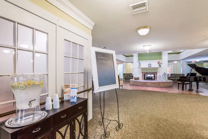 Interior view of a senior living facility common area with a water dispenser and cups on a dark wooden sideboard near glass-paneled doors. A chalkboard sign on an easel reads 'Welcome to Elmcroft.' The room features a carpeted floor, a brick fireplace with decorative items on the mantel, seating areas with armchairs, and a grand piano in the background.