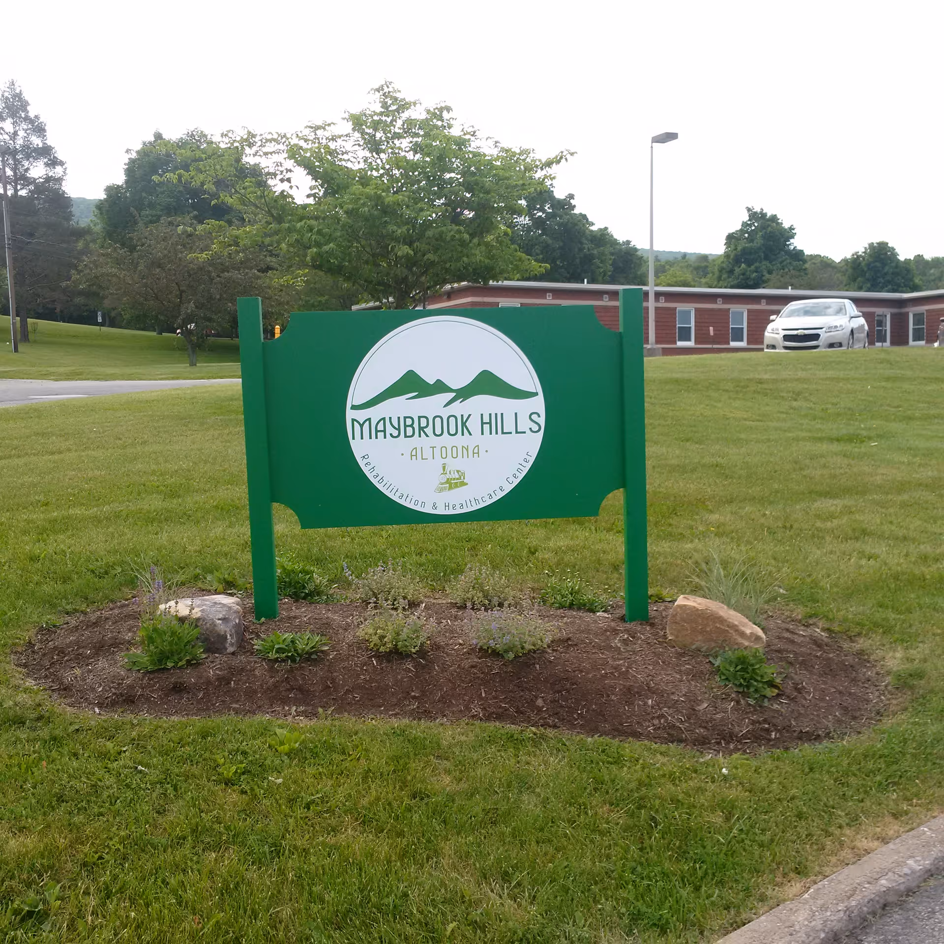 Green sign for Maybrook Hills Rehabilitation and Healthcare Center in a landscaped grassy area with small plants and rocks, with a building and a white car in the background.
