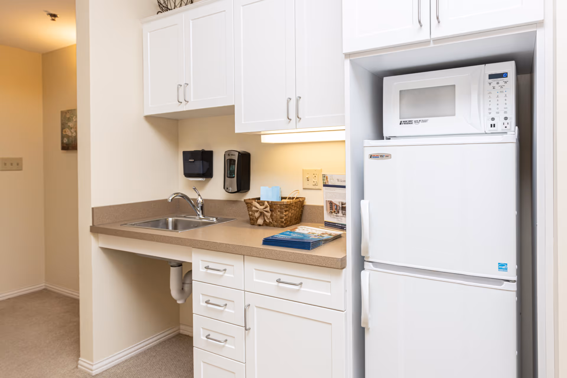 Small kitchenette with a sink, countertop and white cabinets, a microwave atop a refrigerator, and a basket with brochures.
