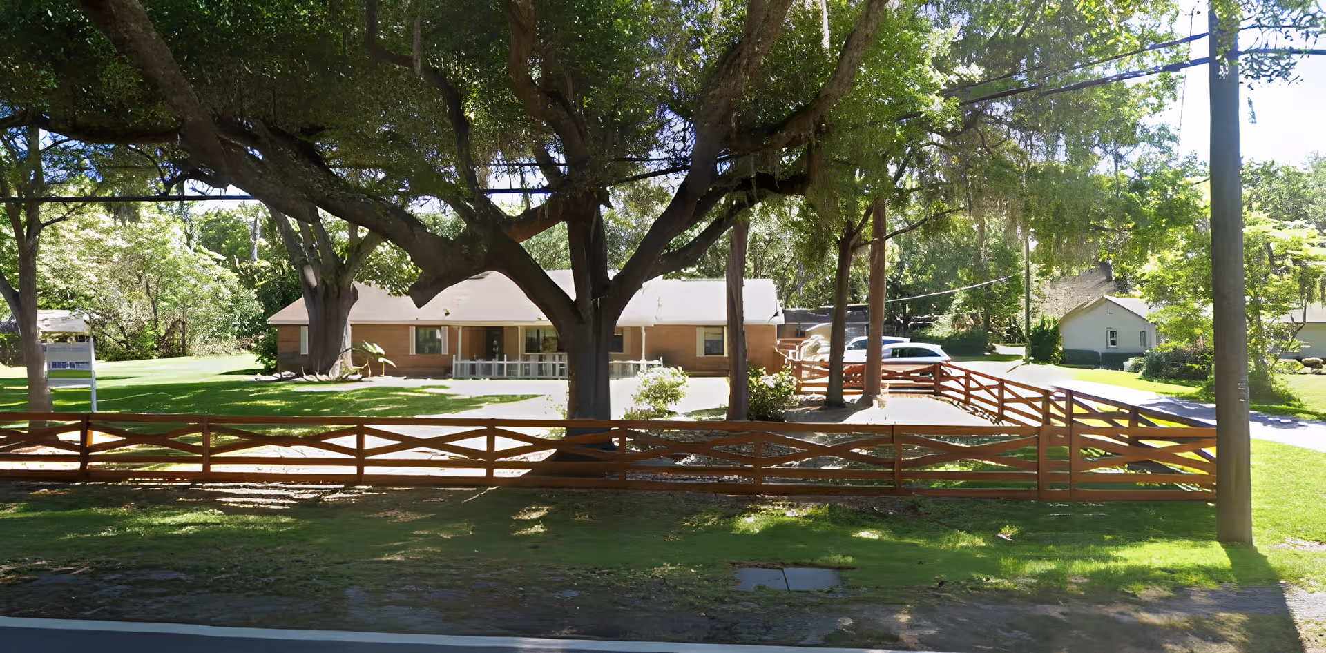 A single-story building with a light-colored roof and brick exterior, surrounded by a wooden fence and large trees providing shade. There is a driveway with parked cars and a well-maintained grassy area with some bushes and smaller trees.