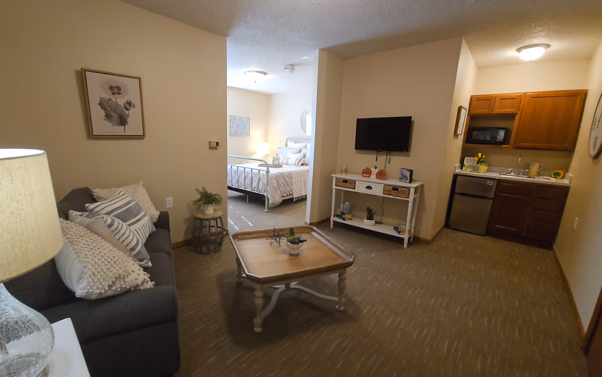 Interior view of an assisted living apartment showing a living room with a gray sofa adorned with multiple pillows, a wooden coffee table with small plants, a TV mounted on the wall above a white console table with decorative items, and a kitchenette with wooden cabinets, a microwave, a small refrigerator, and a sink. In the background, a bedroom with a bed, pillows, and a lamp is visible.