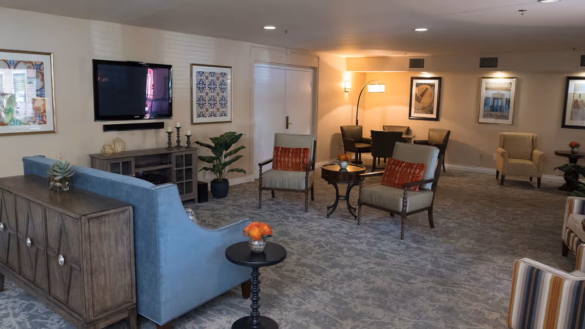 A cozy living room area in a senior living facility featuring a blue sofa, two armchairs with orange cushions, a small round table with a flower vase, a wall-mounted flat-screen TV, framed artwork on the walls, and a carpeted floor. Additional seating and a floor lamp are visible in the background.