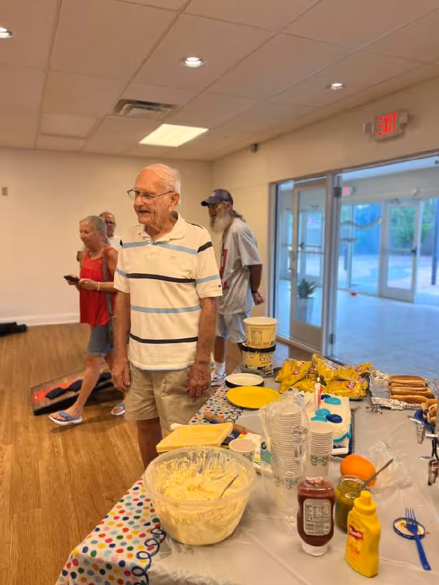 An elderly man in a white and blue striped polo shirt and beige shorts stands near a table with food items including chips, hot dogs, condiments, and a large bowl of salad in a senior living facility. Other seniors are in the background, and the room has wooden flooring and large glass doors leading outside.