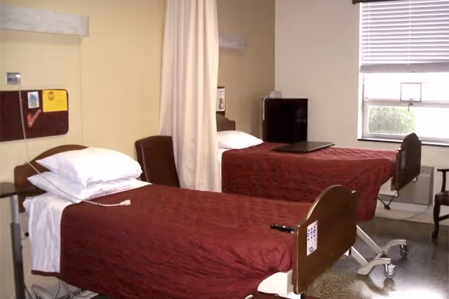 A room with two hospital-style beds covered with maroon bedspreads, each with white pillows. A privacy curtain hangs between the beds. There is a window with blinds partially open, a small refrigerator, and a chair near the window. The floor appears to be polished and the walls are light-colored.