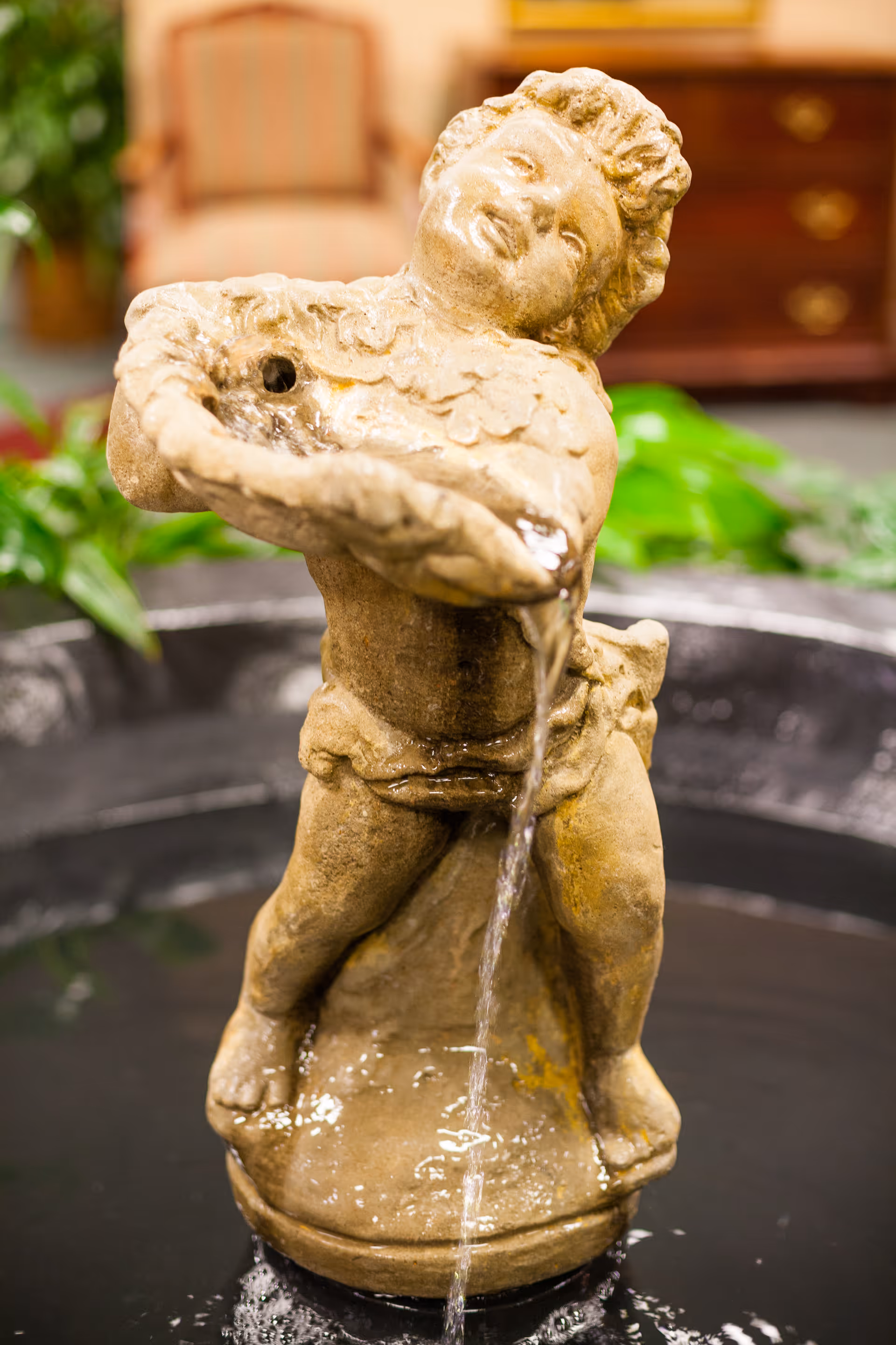 A stone cherub fountain pouring water into a basin with indoor plants and furniture blurred in the background.