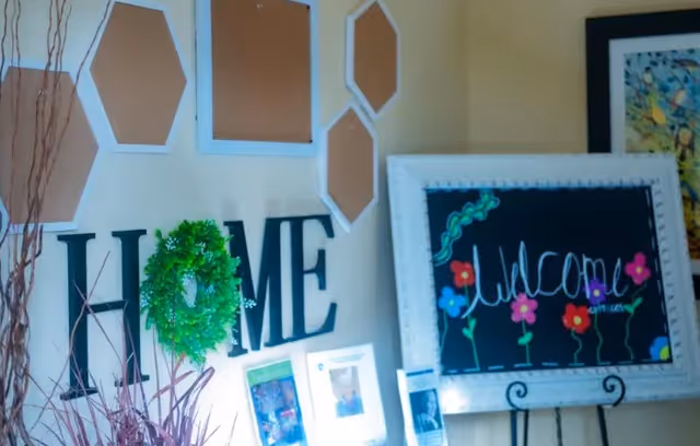 Interior wall decoration featuring the word 'HOME' with a green wreath replacing the letter O, several hexagonal cork boards, and a framed chalkboard sign with colorful flowers and the word 'Welcome' written on it.