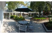Sunny outdoor courtyard with round tables, chairs and umbrellas surrounded by walkways, planters and a single-story building.