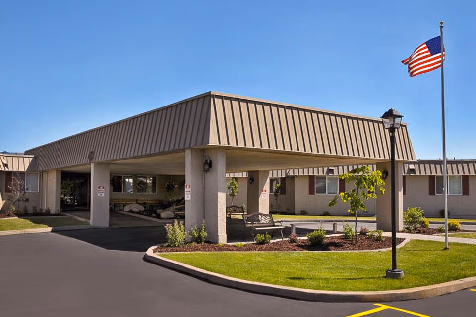 Covered entrance canopy and landscaped circular driveway of a single-story senior living facility with an American flag.