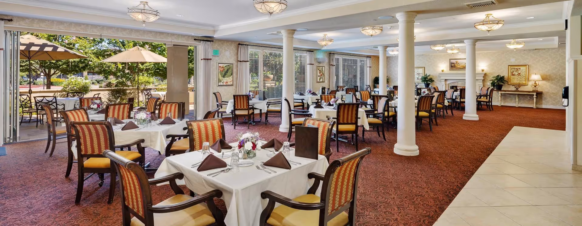 A spacious dining room with multiple tables covered in white tablecloths, each set with brown napkins, glassware, and floral centerpieces. The room features large windows and an open wall leading to an outdoor patio area with tables and umbrellas. The interior has patterned carpet, white columns, and soft lighting fixtures on the ceiling.