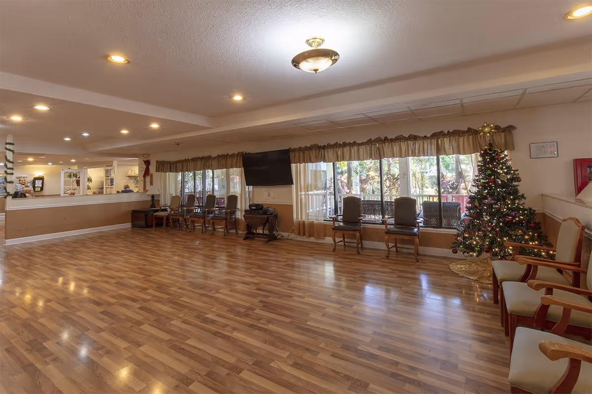 A spacious room with wooden flooring and several chairs arranged along the walls. There is a decorated Christmas tree on the right side near a window with curtains. A flat-screen TV is mounted on the wall above a small table. The room is well-lit with ceiling lights and has large windows showing greenery outside.