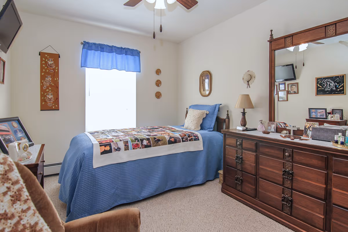 Sunlit bedroom with a single bed in a blue bedspread, a large wooden dresser with mirror, and wall decorations.