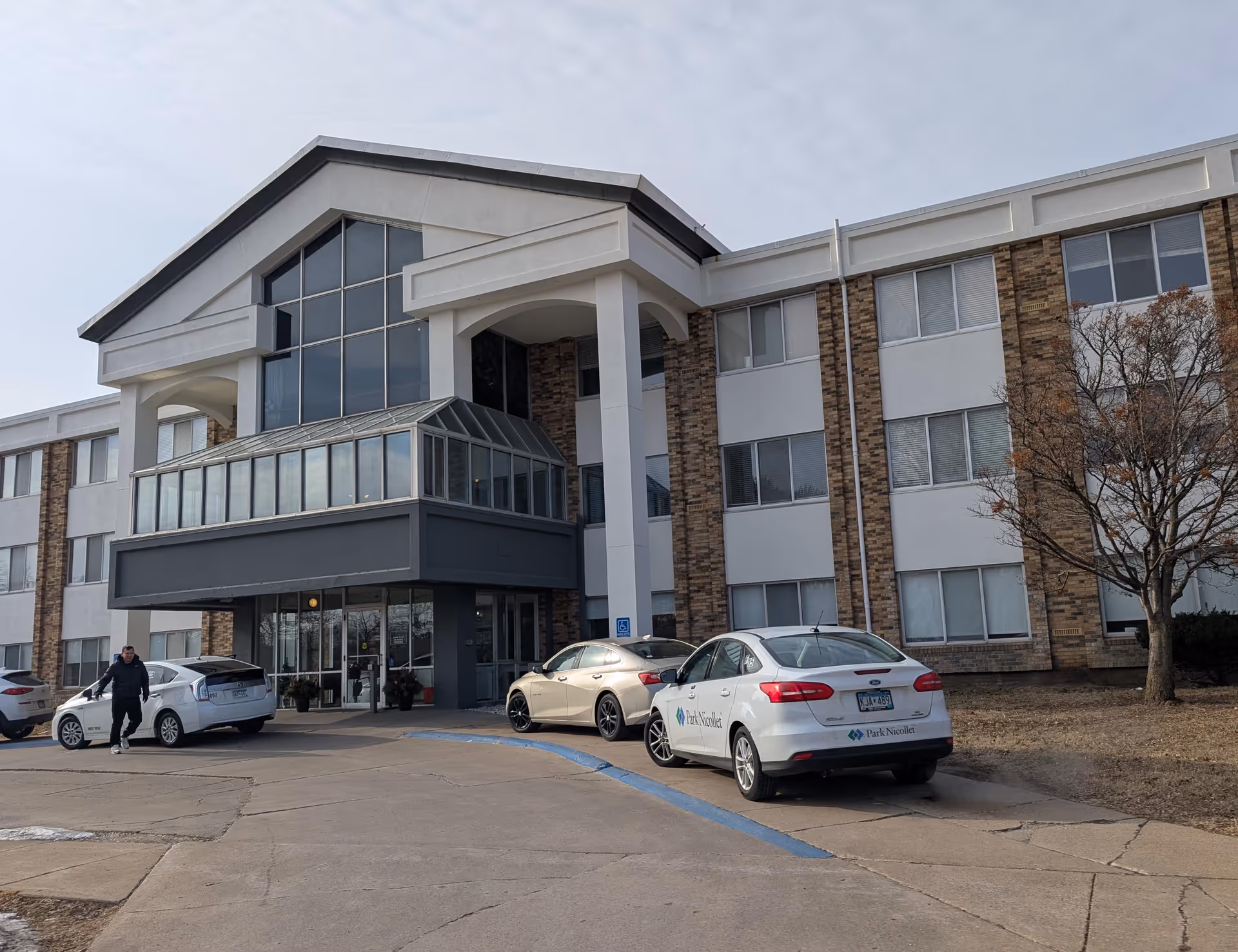Front entrance of a three-story senior living building with a glass atrium and cars parked in the driveway.