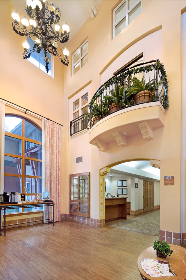Interior view of a senior living facility lobby with high ceilings, a large decorative chandelier, a balcony with potted plants, a window with pink curtains, a table with refreshments, and a reception desk in the background.