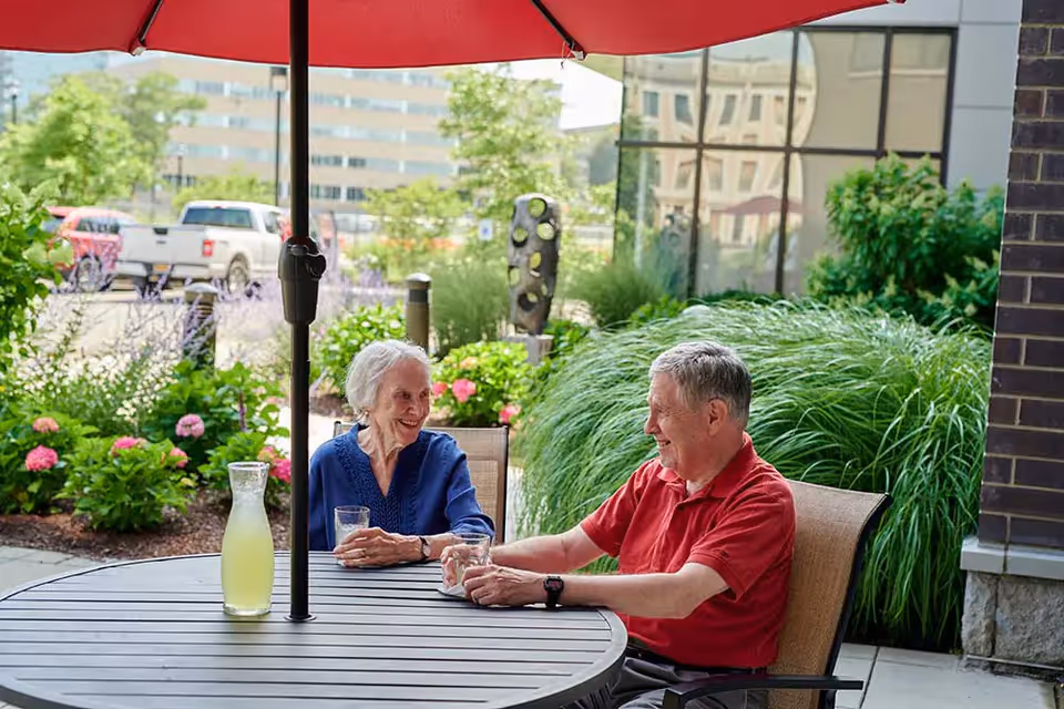 Two elderly people sitting at a round outdoor table under a red umbrella, enjoying drinks and smiling at each other. The background shows greenery, flowers, and a building with reflective windows.