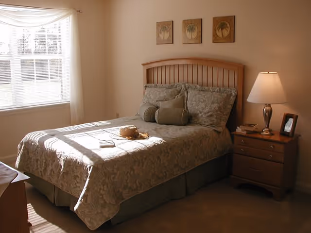 Sunlit bedroom with a made bed, wooden headboard, bedside table with a lamp, and a window with blinds.