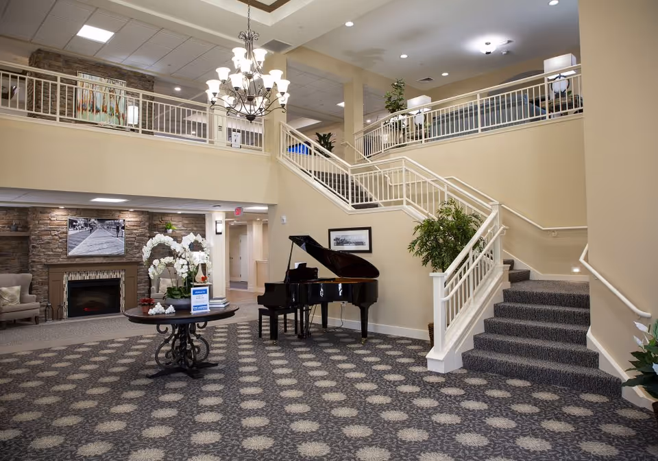 Spacious senior living facility common area with a patterned carpet, grand piano, round table with white orchids, and a stone fireplace with a black and white photo above it. There is a staircase leading to an upper level with seating areas and plants, under warm lighting and a chandelier.