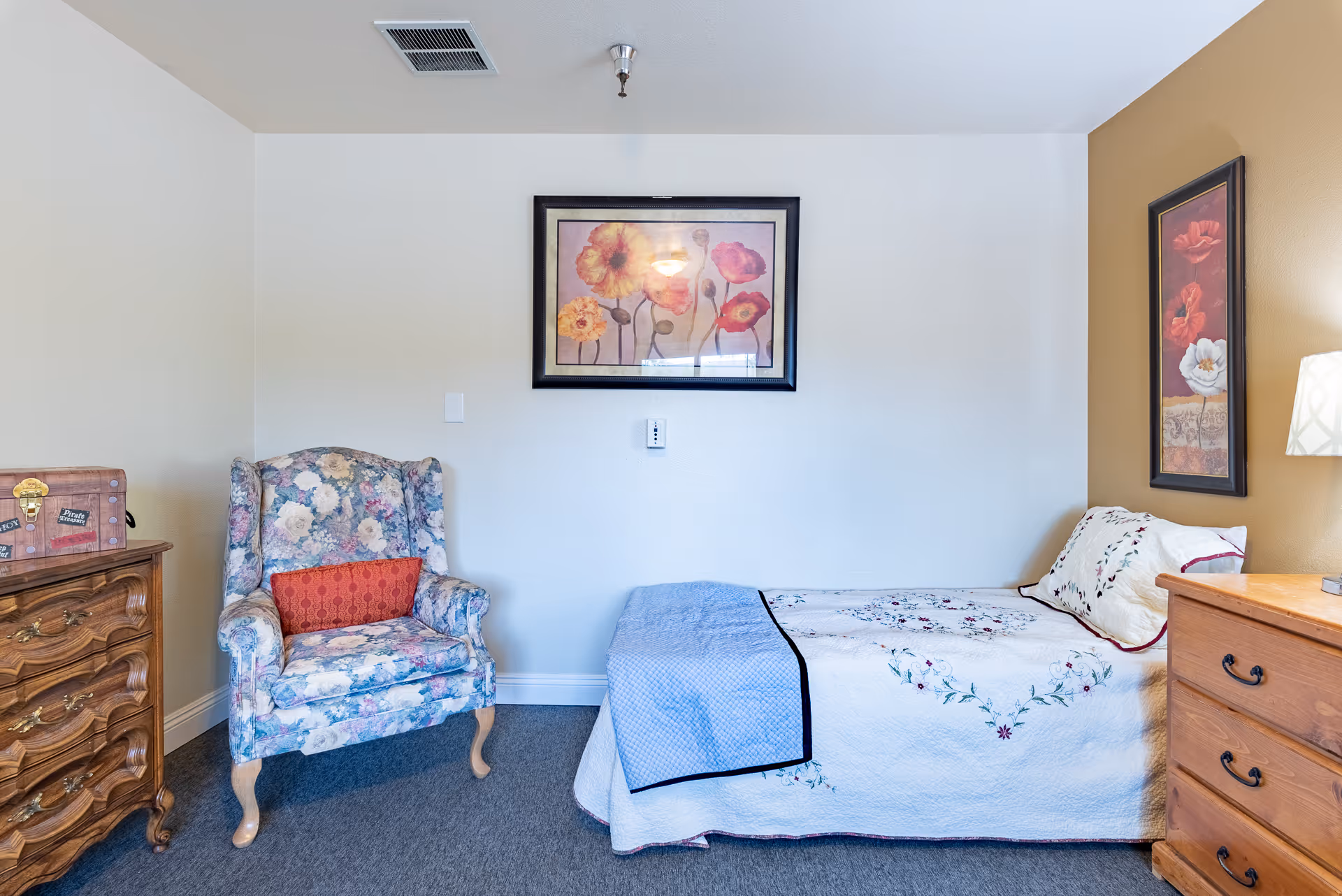 A cozy bedroom with a single bed covered in a white quilt with floral embroidery and a light blue blanket. Next to the bed is a wooden dresser with a lamp on top. A floral upholstered armchair with a red cushion sits beside another wooden dresser with a decorative chest on it. Two framed floral paintings hang on the walls, one above the bed and one on the adjacent wall.