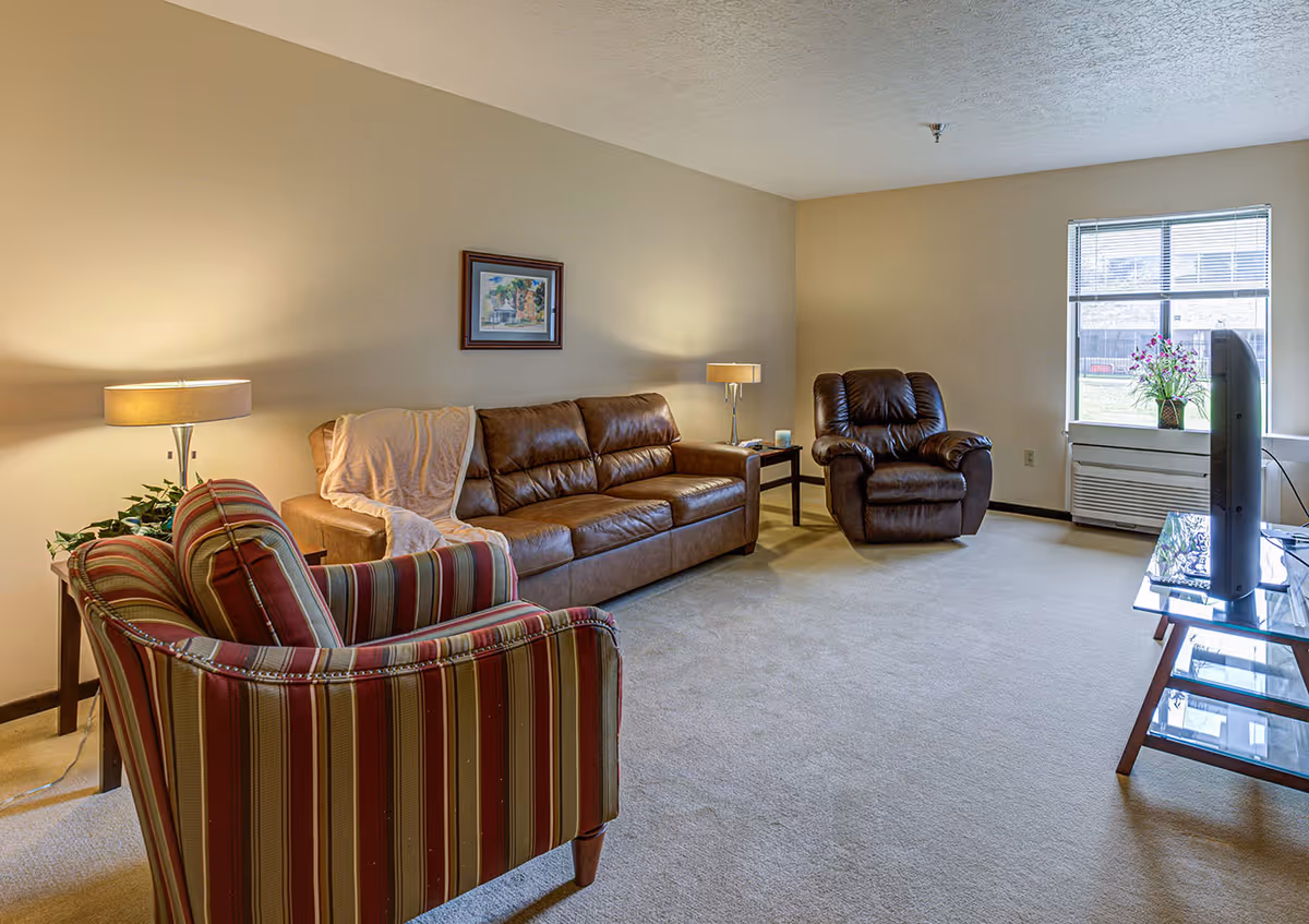 A cozy living room with a brown leather sofa, a striped armchair, and a brown recliner. There are two side tables with lamps on either side of the sofa, a framed picture on the wall, a window with blinds letting in natural light, and a TV on a glass stand.