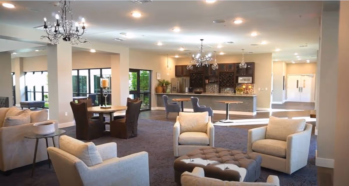 A spacious and well-lit common area in a senior living facility featuring comfortable armchairs arranged around a tufted ottoman, a round table with chairs, and a kitchen area with bar stools and cabinetry in the background. The room has chandeliers and large windows letting in natural light.
