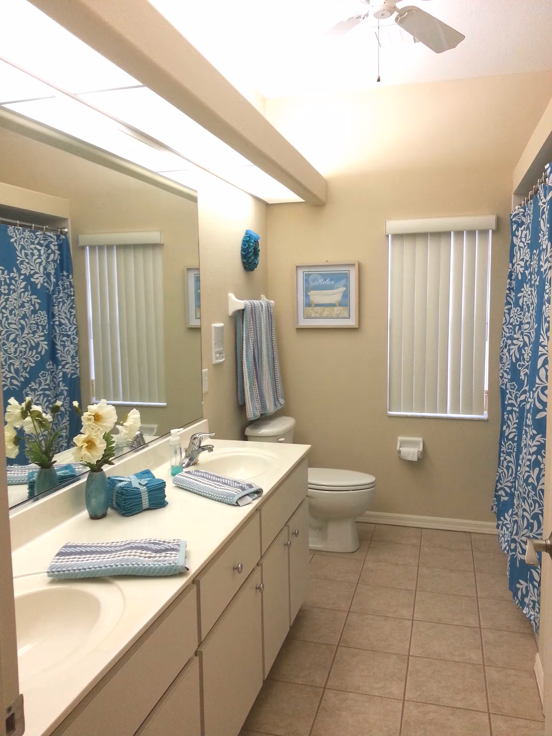 A clean and bright bathroom with a double sink vanity topped with folded blue and white towels and a vase of white flowers. There is a large mirror above the sinks, a toilet next to a window with vertical blinds, and a shower with a blue and white patterned curtain. The walls are light beige and the floor is tiled.