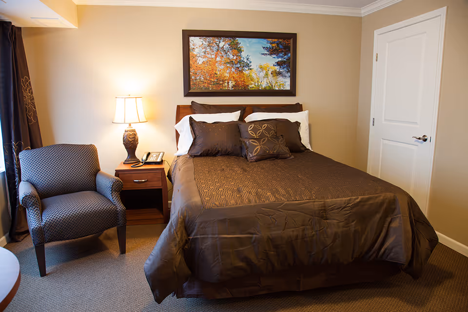 A cozy bedroom with a neatly made bed featuring brown bedding and multiple pillows. To the left of the bed is a wooden nightstand with a decorative lamp and a telephone. Next to the nightstand is a patterned armchair. A framed picture of an autumn landscape hangs on the beige wall above the bed. A white door is visible on the right side of the room.
